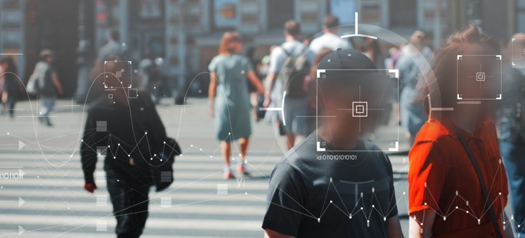 Imagem mostra pessoas andando em uma rua, em cima de uma faixa de pedestres; seus rostos estão borrados e, na frente, há uma ilustração de um leitor de reconhecimento facial, ilustrando uma das possibilidades que a tecnologia de inteligência artificial pode fazer