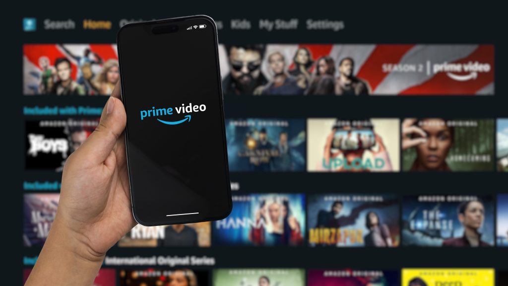 Imagem mostra um smartphone com a logomarca do Amazon Prime Video à frente de um fundo com as séries e filmes do serviço de streaming