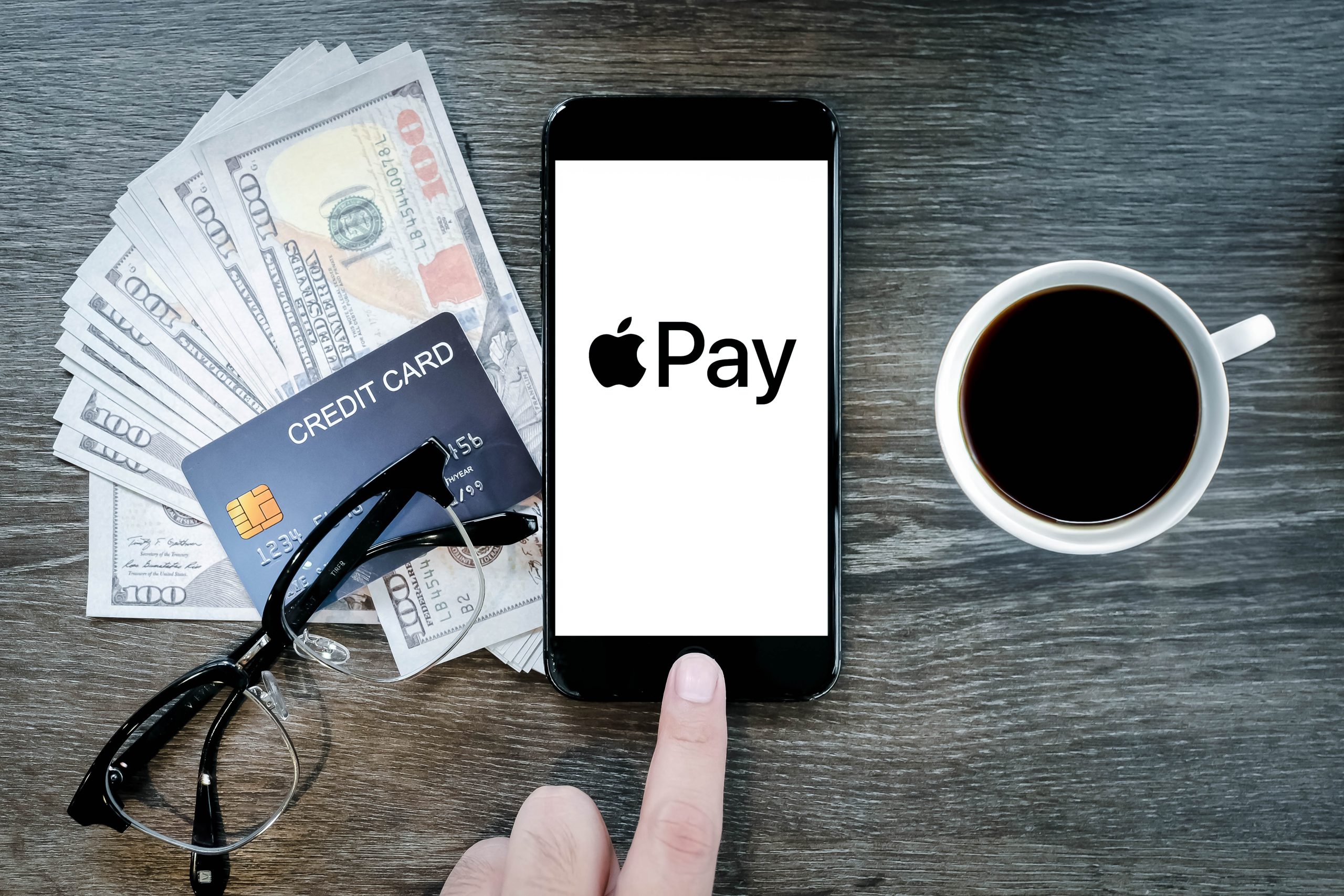 Imagem mostra tela do Apple Pay aberta em um smartphone, próxima a um copo com café, um par de óculos, notas variadas de dólares e um cartão de crédito