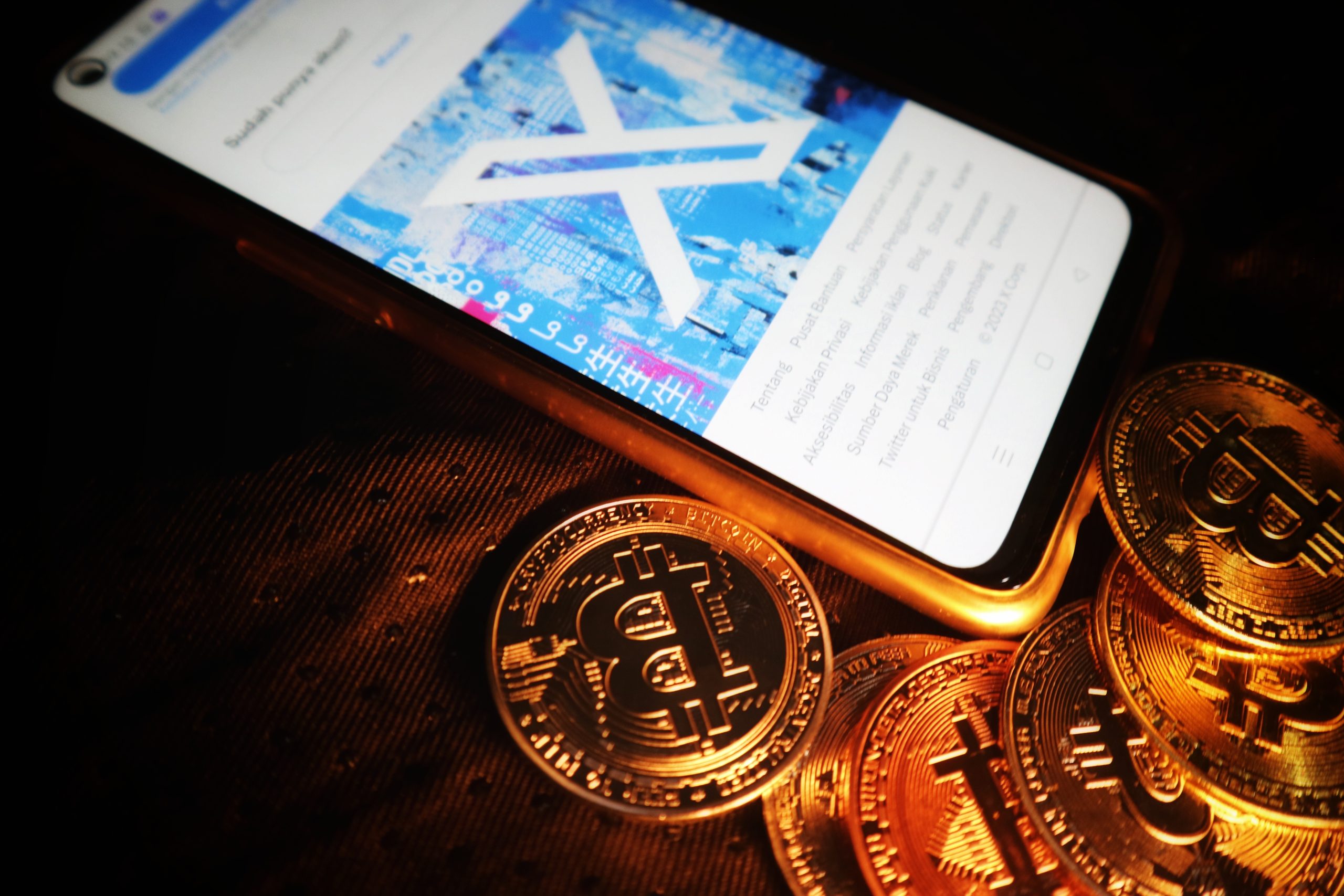 Imagem mostra o X na tela de um smartphone em meio a várias moedas com o símbolo do bitcoin, simbolizando recursos de fintechs
