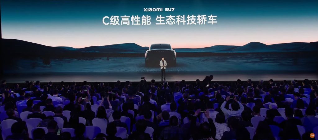 Imagem mostra um palco onde está o CEO da Xiaomi durante a apresentação do carro elétrico da empresa, o SU7. ao fundo há uma tela com uma imagem do carro e, à frente, a plateia.