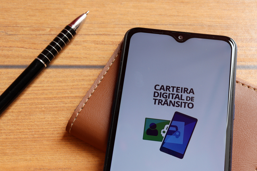 Em cima de uma superfície de madeira estão uma caneta e uma carteira de couro; em cima da carteira está um smartphone onde dá para ver o logo da Carteira Digital de Trânsito (CDT) na tela, ilustrando o aplicativo da cnh digital brasileira