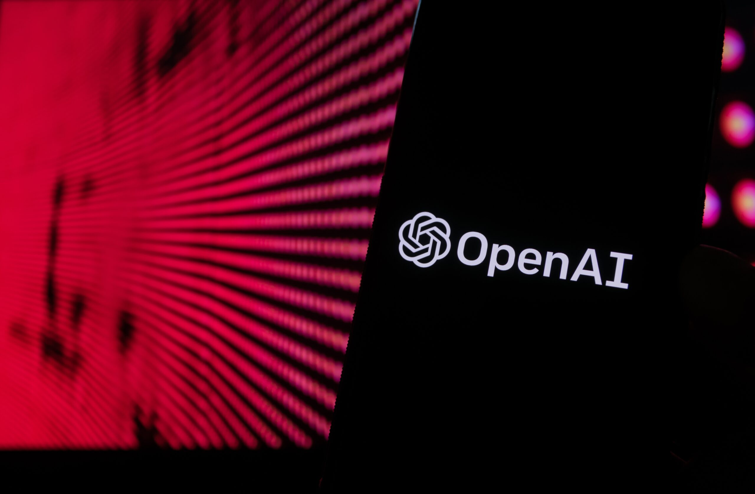 Imagem mostra um smartphone com a logomarca da OpenAI na tela, à frente de um monitor de computador com vários códigos de programação em vermelho